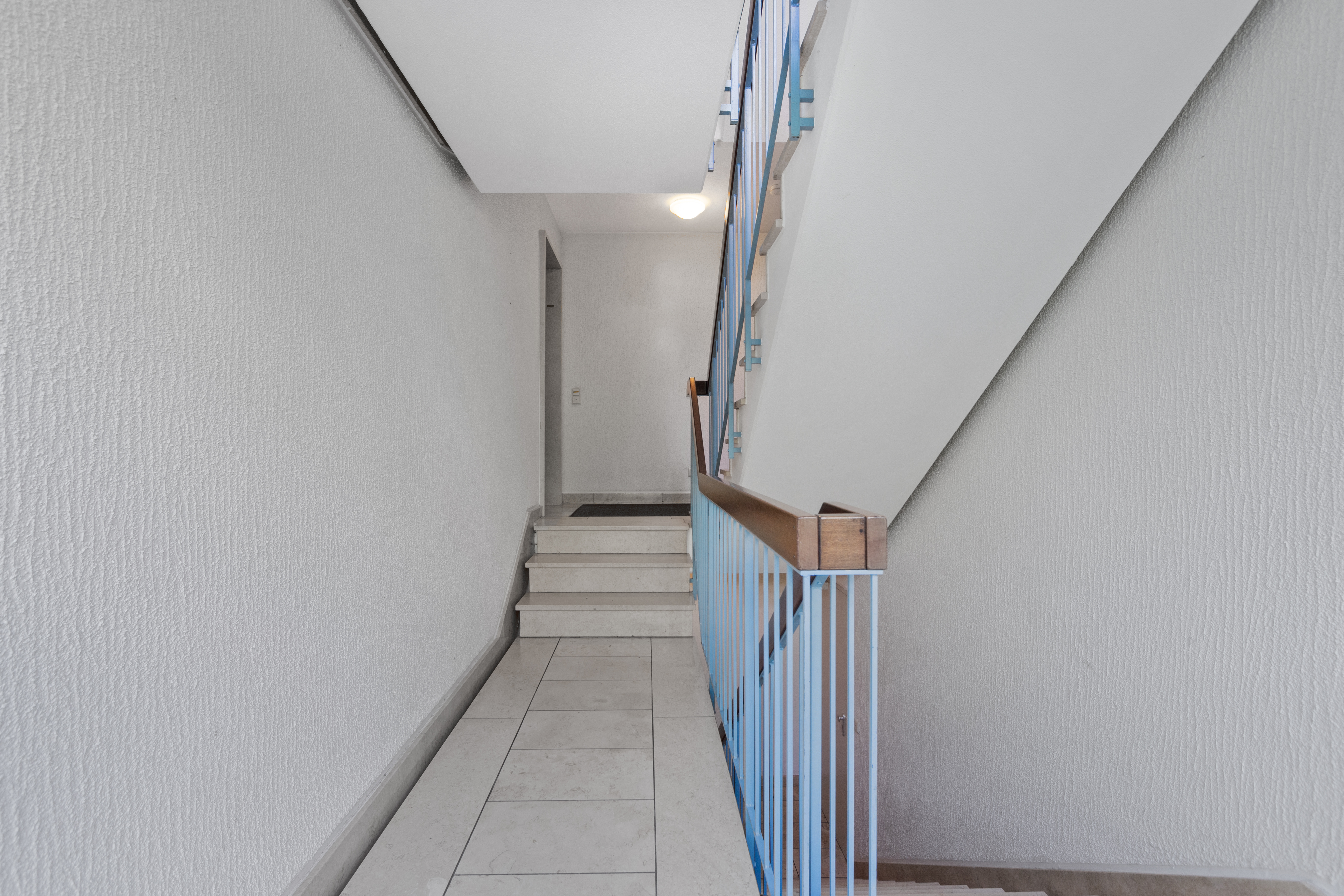 Helles Treppenhaus mit weißen Wänden und blauem Geländer in einem Wohnhaus.