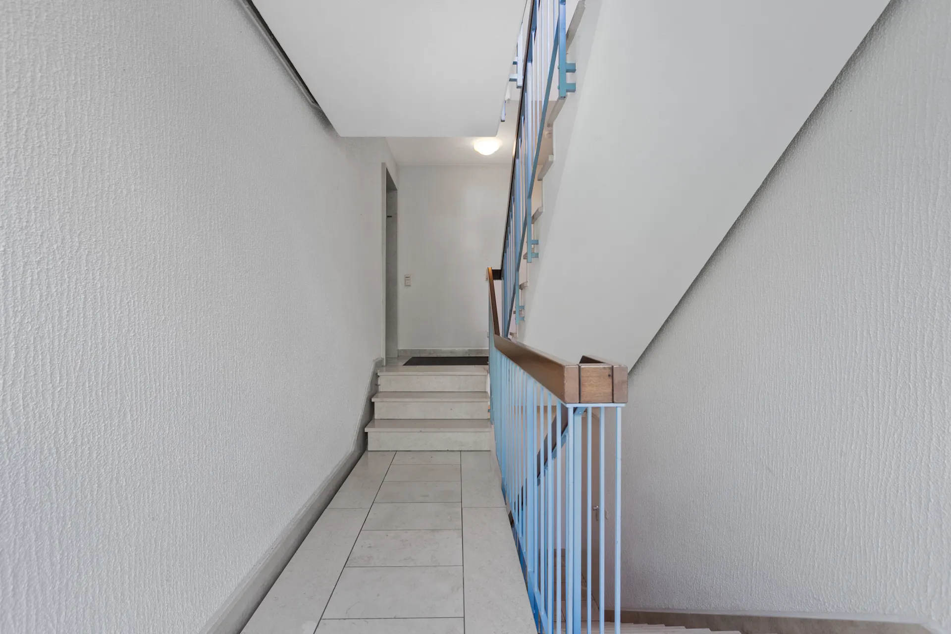 Helles Treppenhaus mit weißen Wänden und blauem Geländer in einem Wohnhaus.