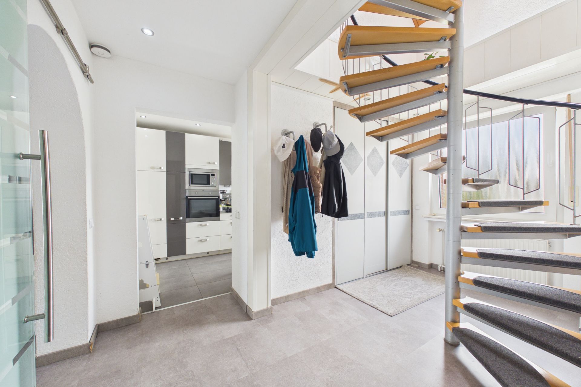 Heller Flur mit Holz-Wendeltreppe, Garderobe und Blick in eine moderne Küche.