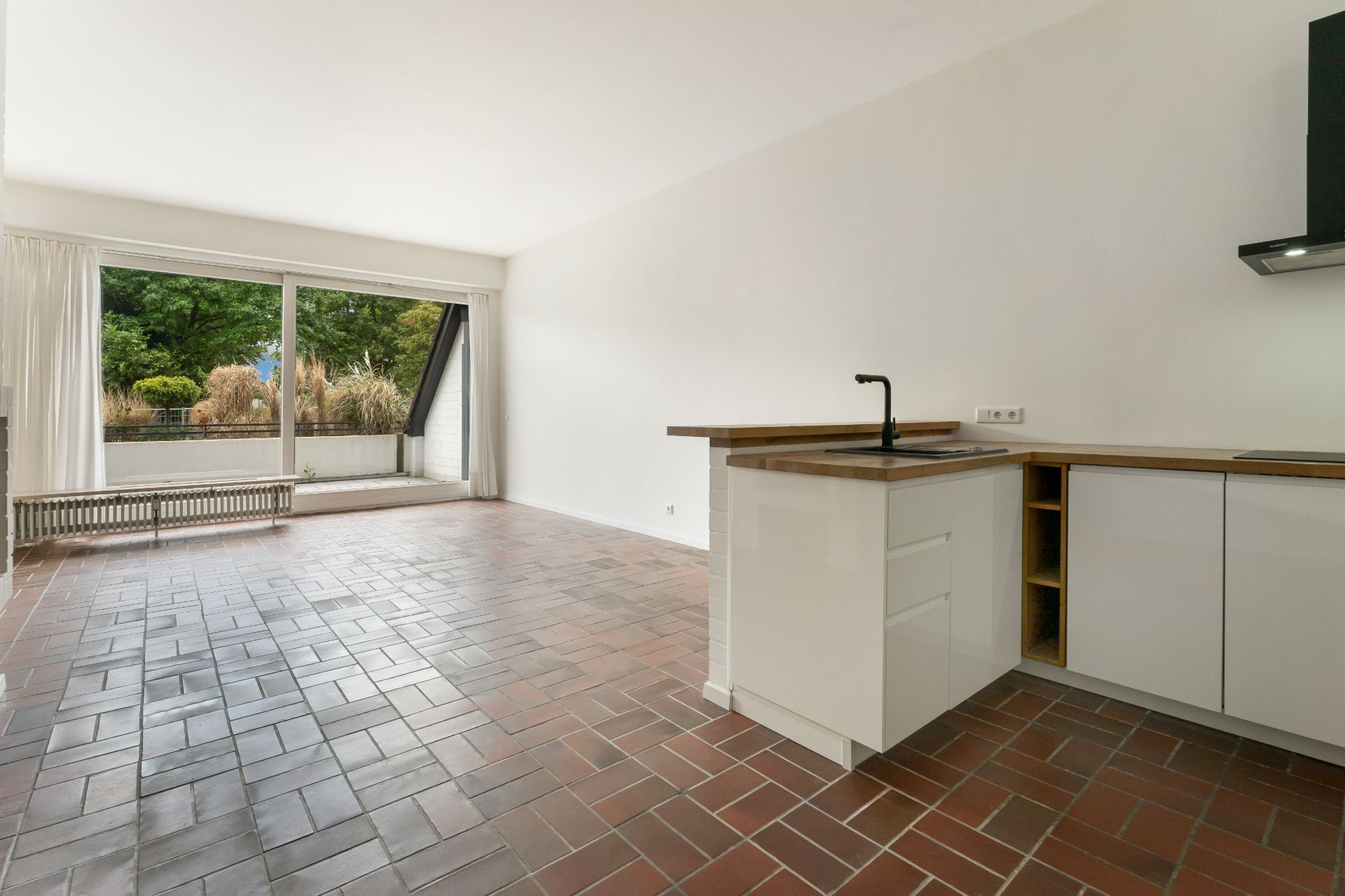 Heller Wohnraum mit offener Küche, Fliesenboden und großen Fenstern zur Terrasse.
