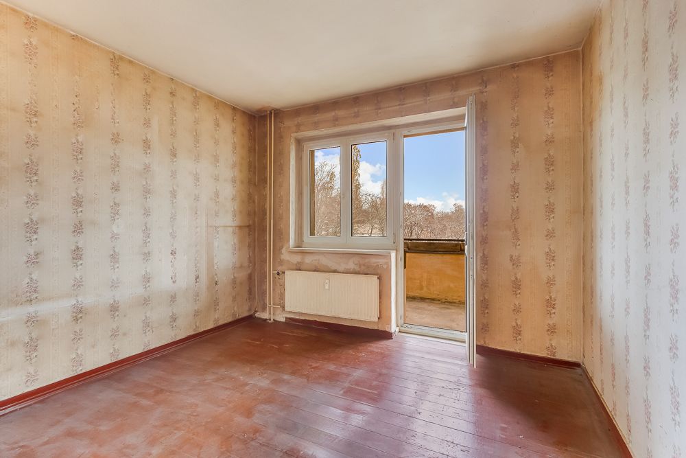 Leeres Zimmer mit Tapeten, Holzboden, Heizkörper und offenem Fenster zur kleinen Balkonfläche.