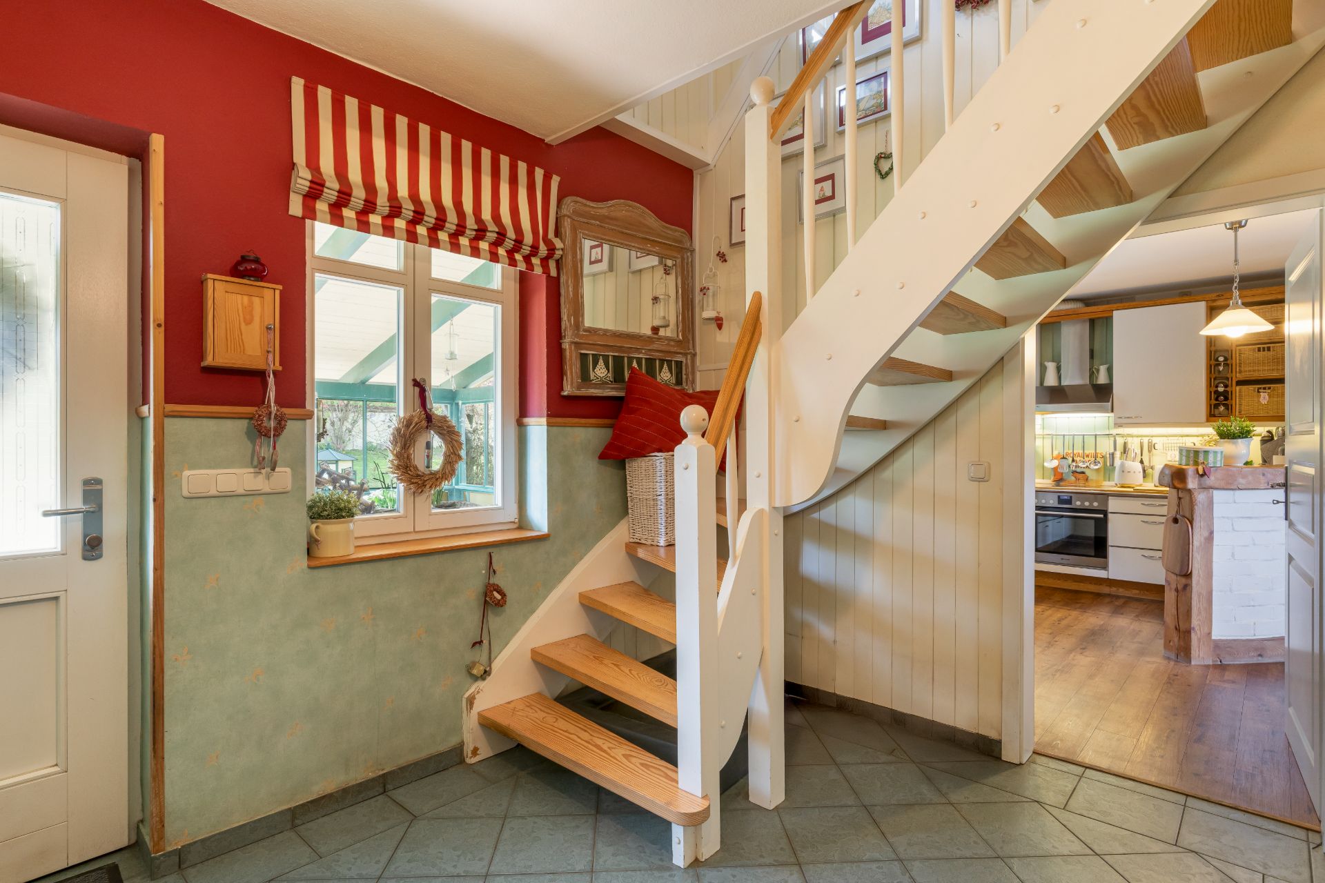 Eingangsbereich mit Treppe, Fenster mit gestreiftem Vorhang und Blick in eine helle Küche.