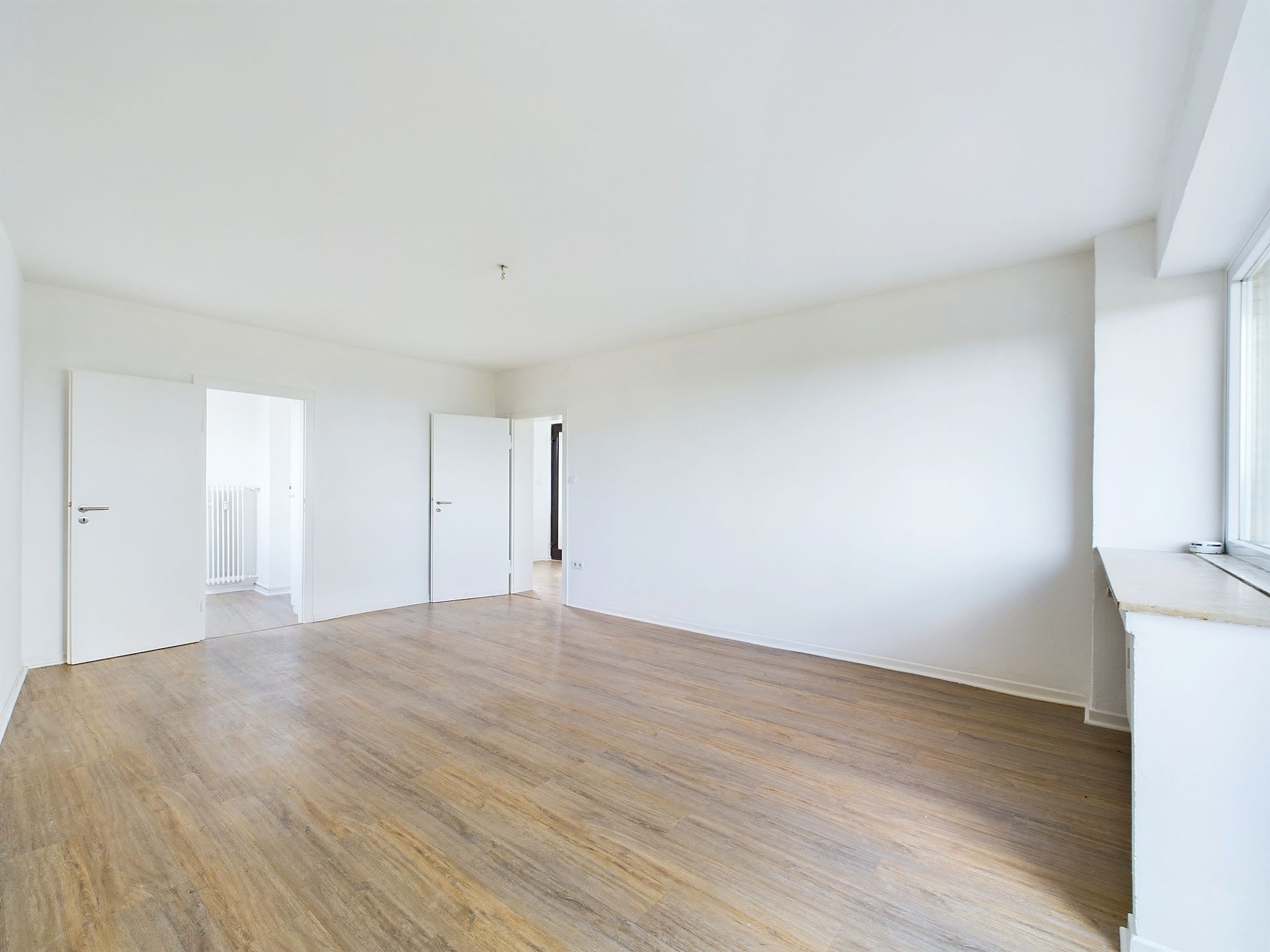 Helles, leeres Zimmer mit Holzboden, weißen Wänden und zwei offenen Türen.