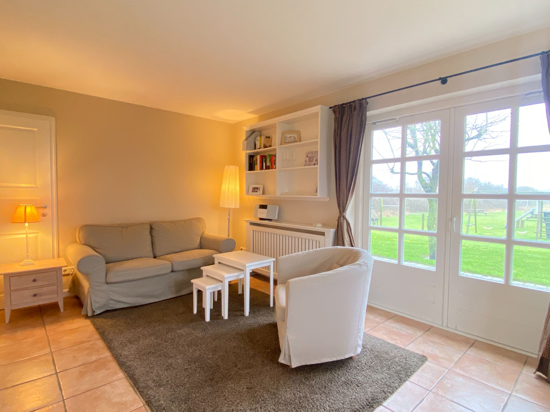 Helles Wohnzimmer mit Sofa, Sessel, kleinen Tischen und großen Fenstern mit Blick auf den Garten.