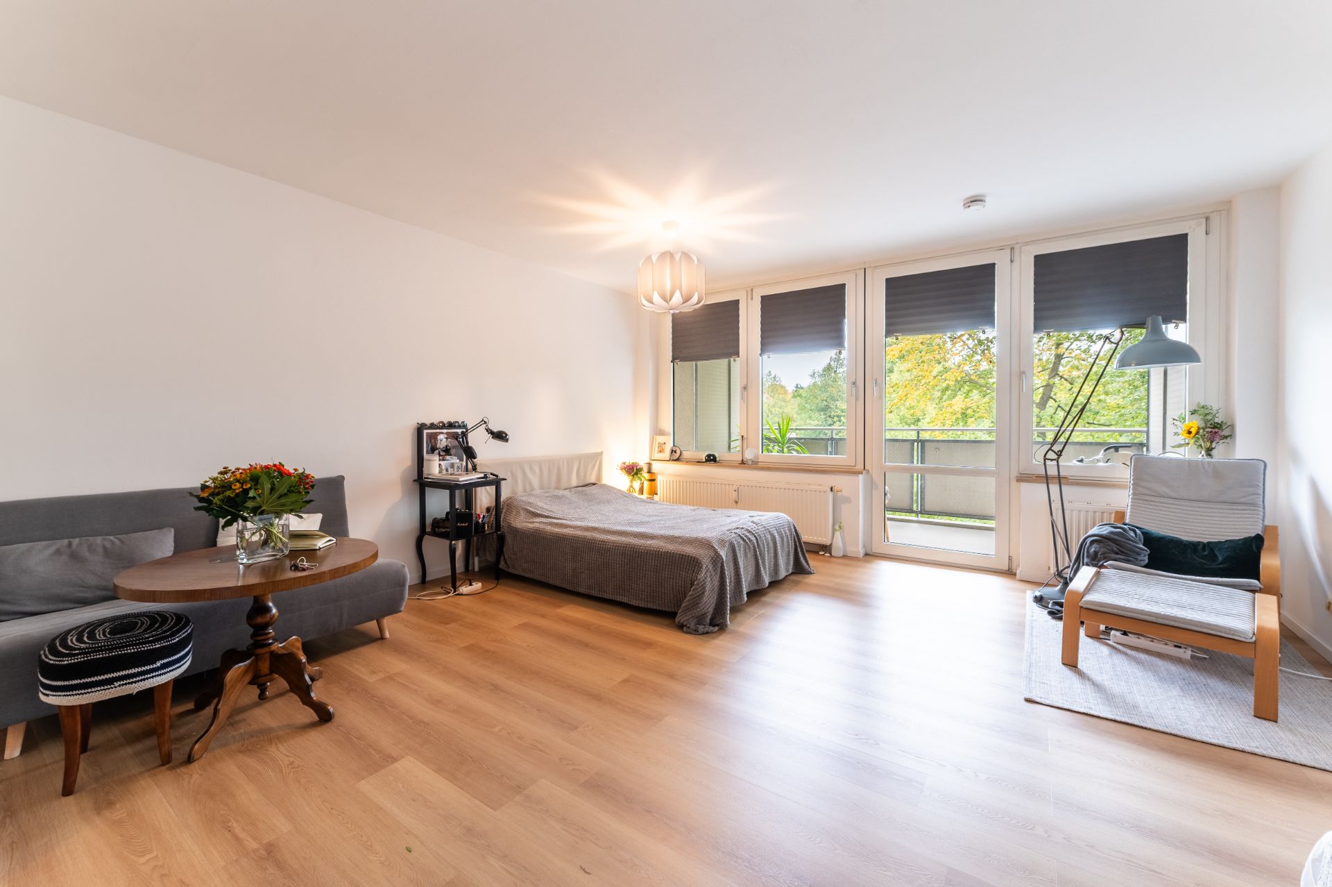 Helles Wohnzimmer mit Bett, Sofa, Sessel, Holzboden und großen Fenstern mit Blick ins Grüne.