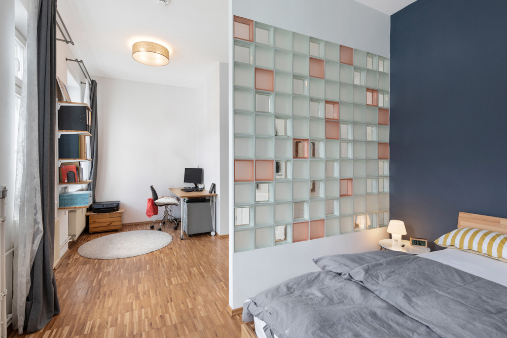 Zimmer mit Arbeitsplatz, Regal und Bett, getrennt durch Glasbausteinwand.
