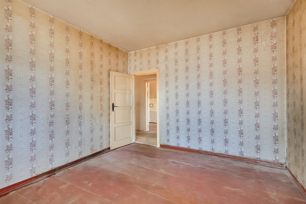 Leeres Zimmer mit gemusterter Tapete, Holzboden und geöffneter weißer Tür.