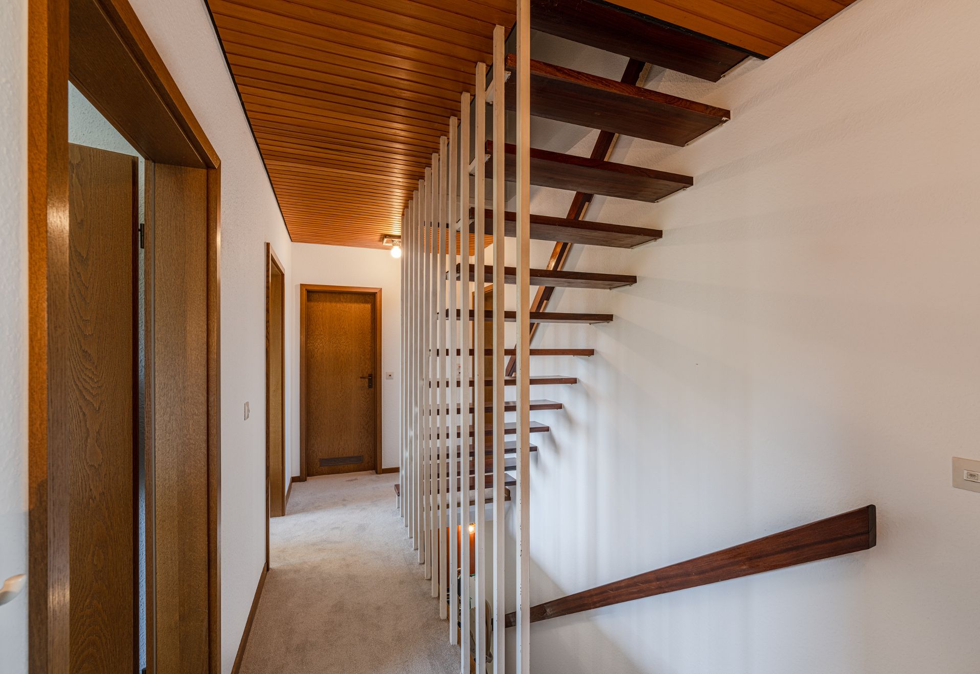 Flur mit Teppichboden, Holzdecke und offener Holztreppe mit weißen Streben.