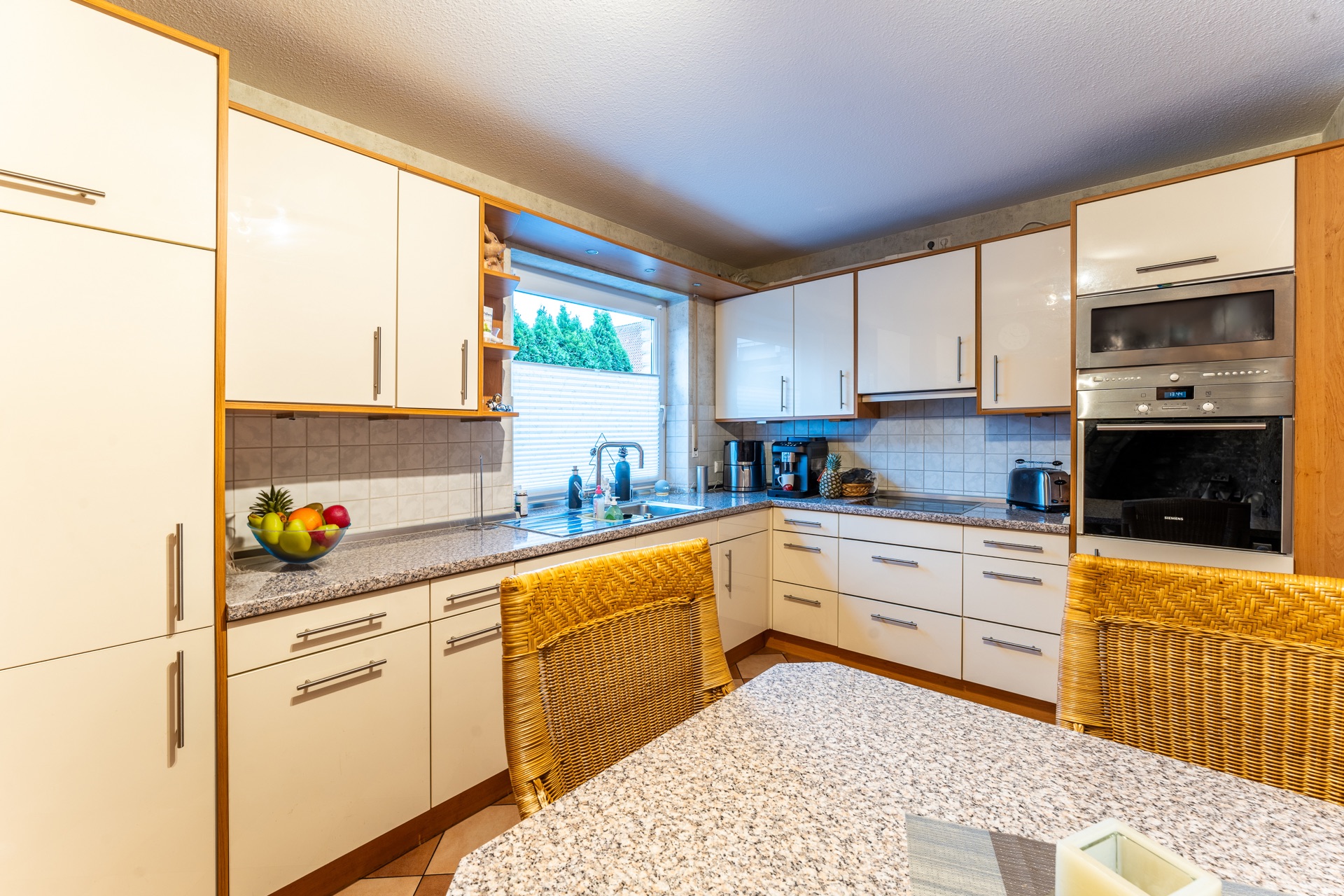 Moderne Küche mit weißen Schränken, Früchteschale, Esstisch und großem Fenster.