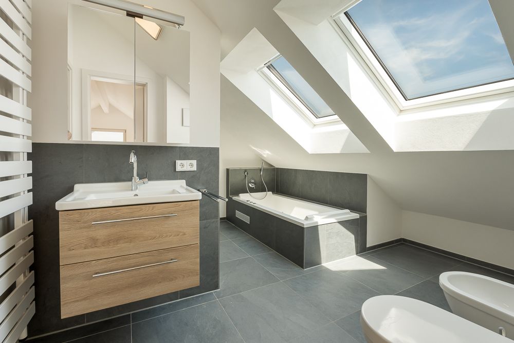 Helles Dachgeschoss-Badezimmer mit Badewanne, Waschbecken, Spiegel und großen Dachfenstern.