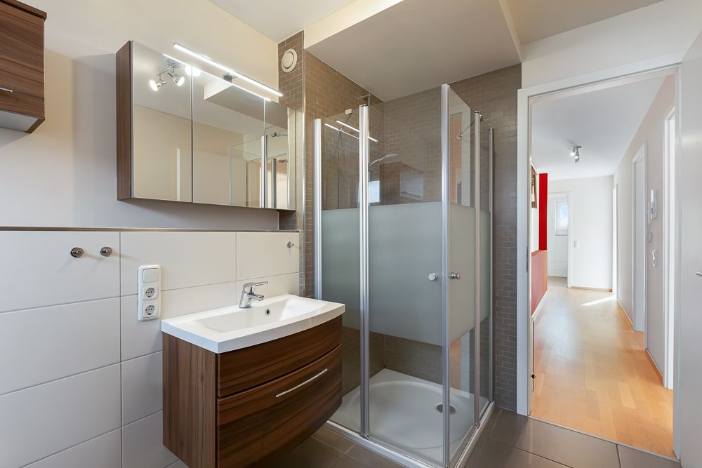Modernes Badezimmer mit Dusche, Waschbecken und Spiegel, angrenzend an einen Flur mit Holzboden.