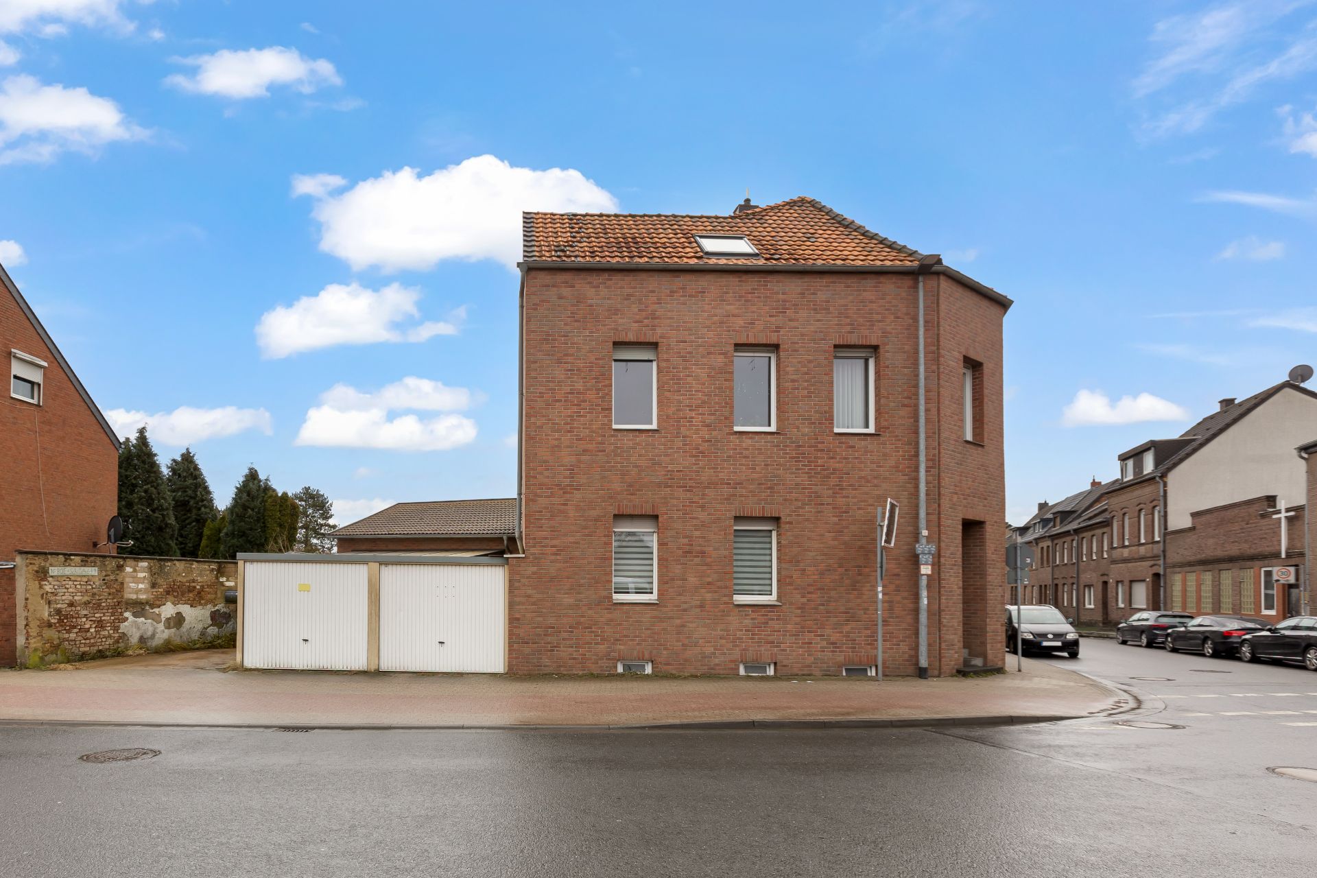 Eckhaus aus rotem Backstein mit mehreren Fenstern, daneben weiße Garage, Straße im Vordergrund.
