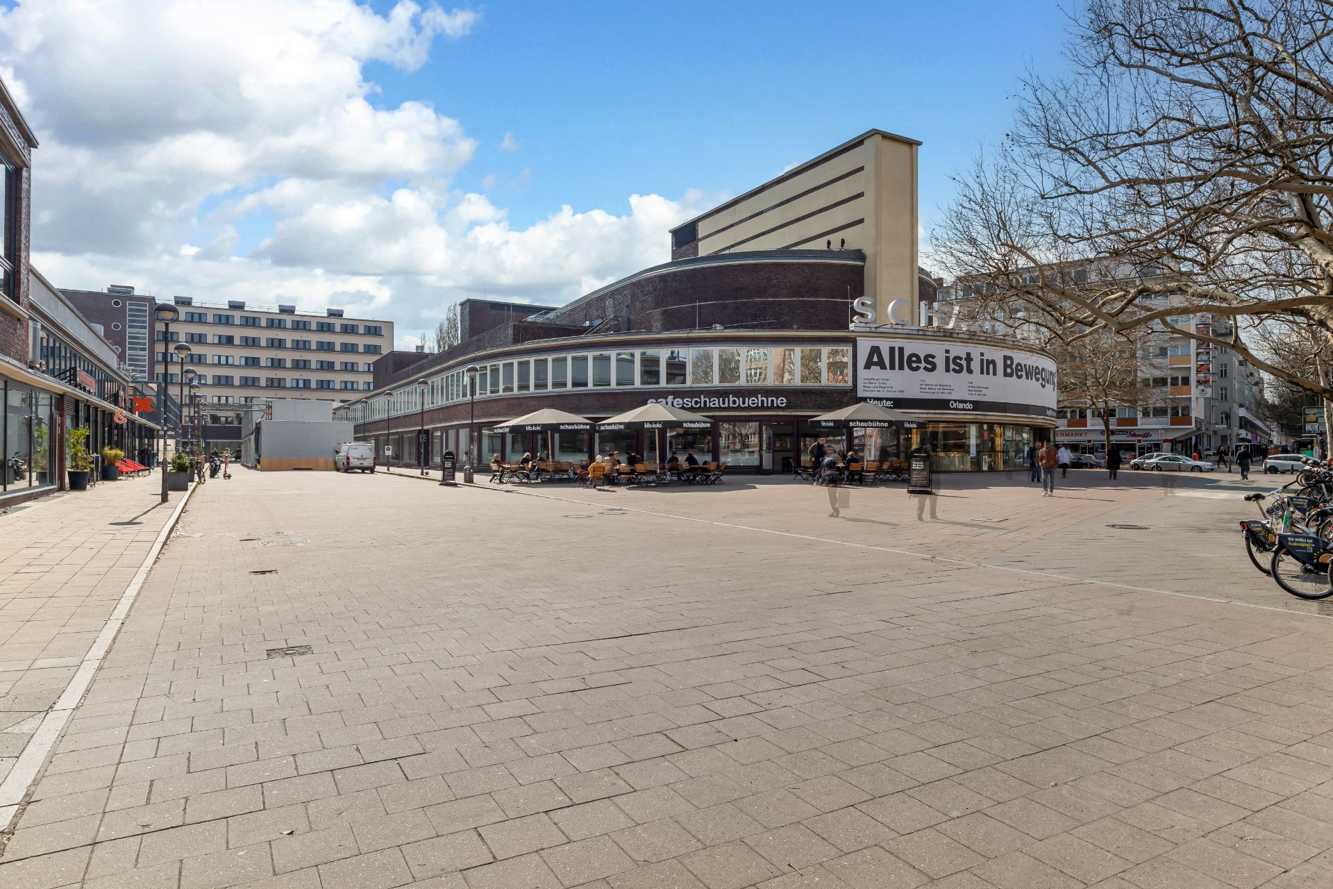 Weitläufiger Platz mit Bäumen, Fahrrädern und einem modernen Gebäude mit großer Fensterfront.