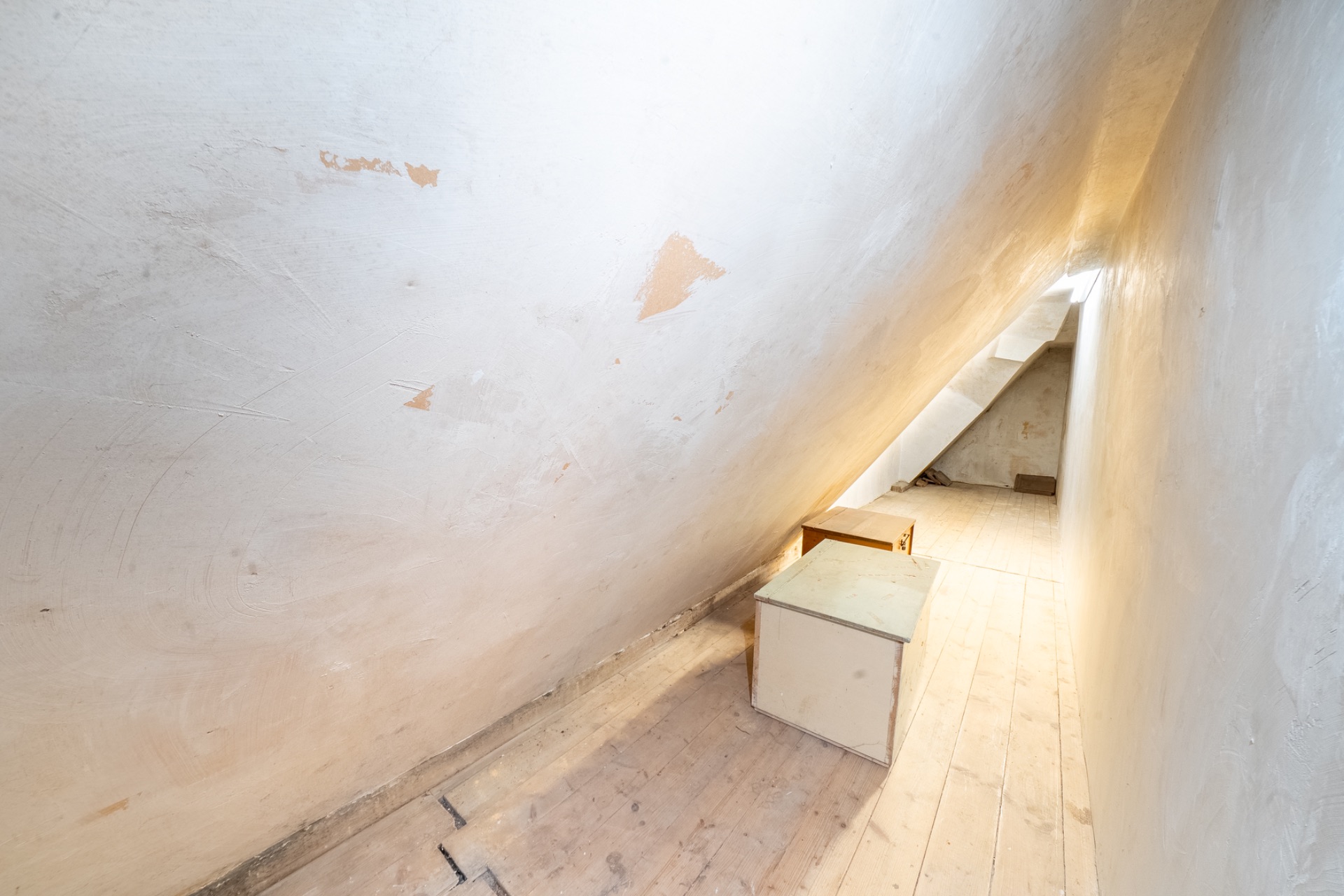 Heller, schmaler Dachboden mit schräger Wand und zwei kleinen Möbelstücken aus Holz.