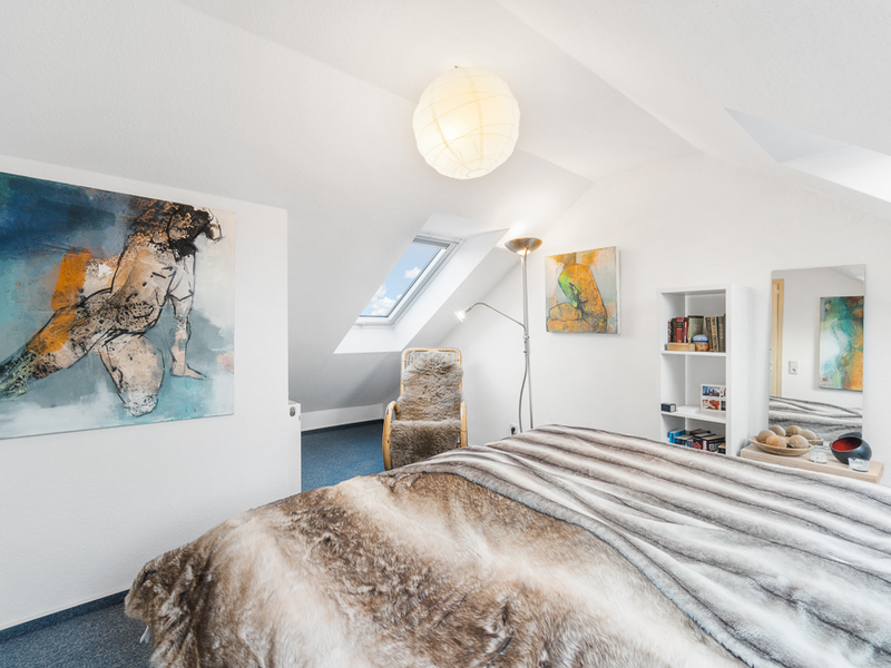 Helles Schlafzimmer mit Felldecke, Kunstwerken, Sessel und Bücherregal unter Dachschrägen.