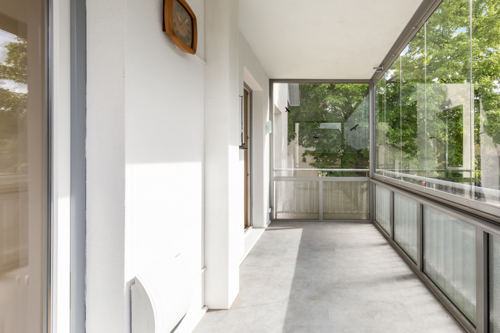 Heller, verglaster Balkon mit Blick auf grüne Bäume und einer Wanduhr.