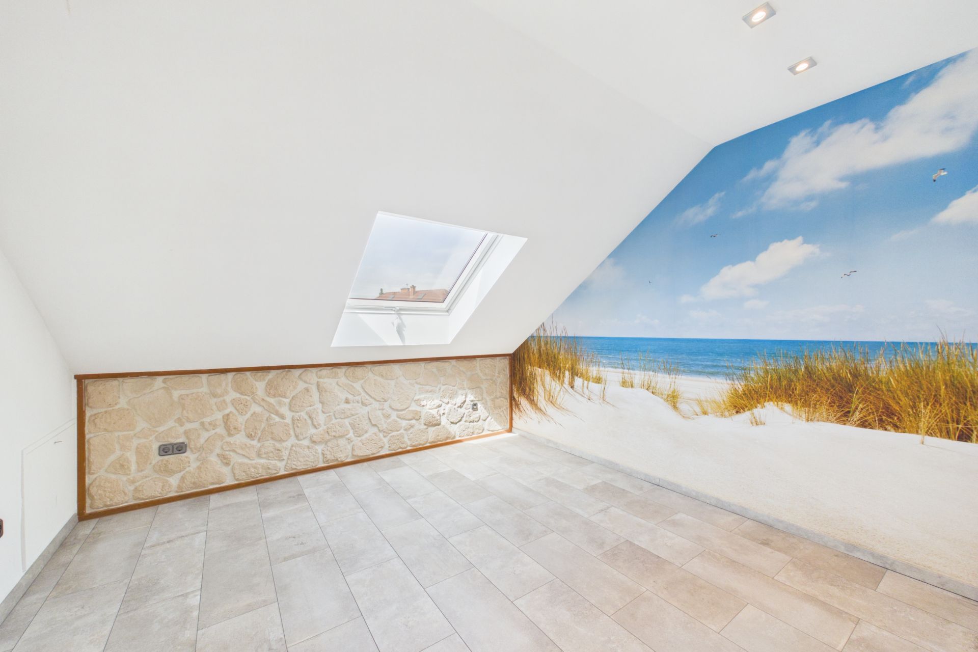 Heller Raum mit Dachfenster und Wandbild eines Strandes mit Meer und Dünen.