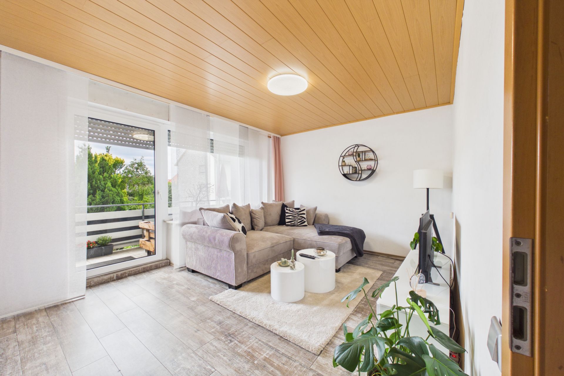 Helles Wohnzimmer mit Ecksofa, Couchtischen, Balkonzugang und Zimmerpflanzen.