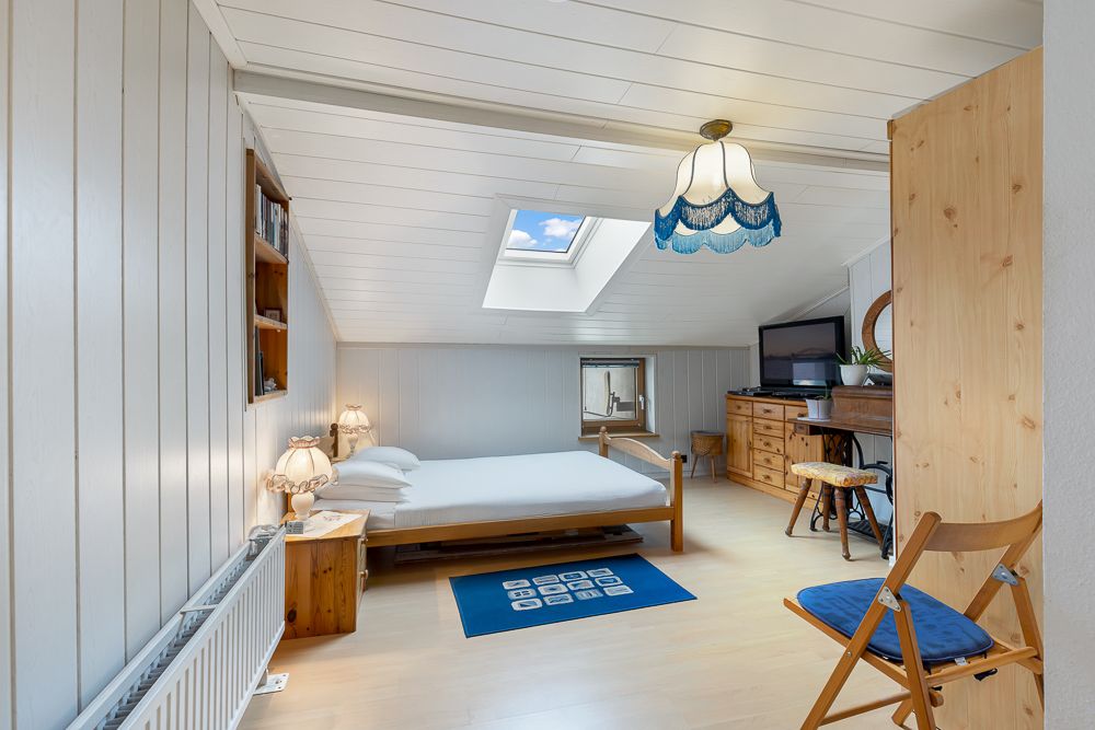 Helles Schlafzimmer mit Dachfenster, Bett, Holzmöbeln und einer blauen Hängelampe.