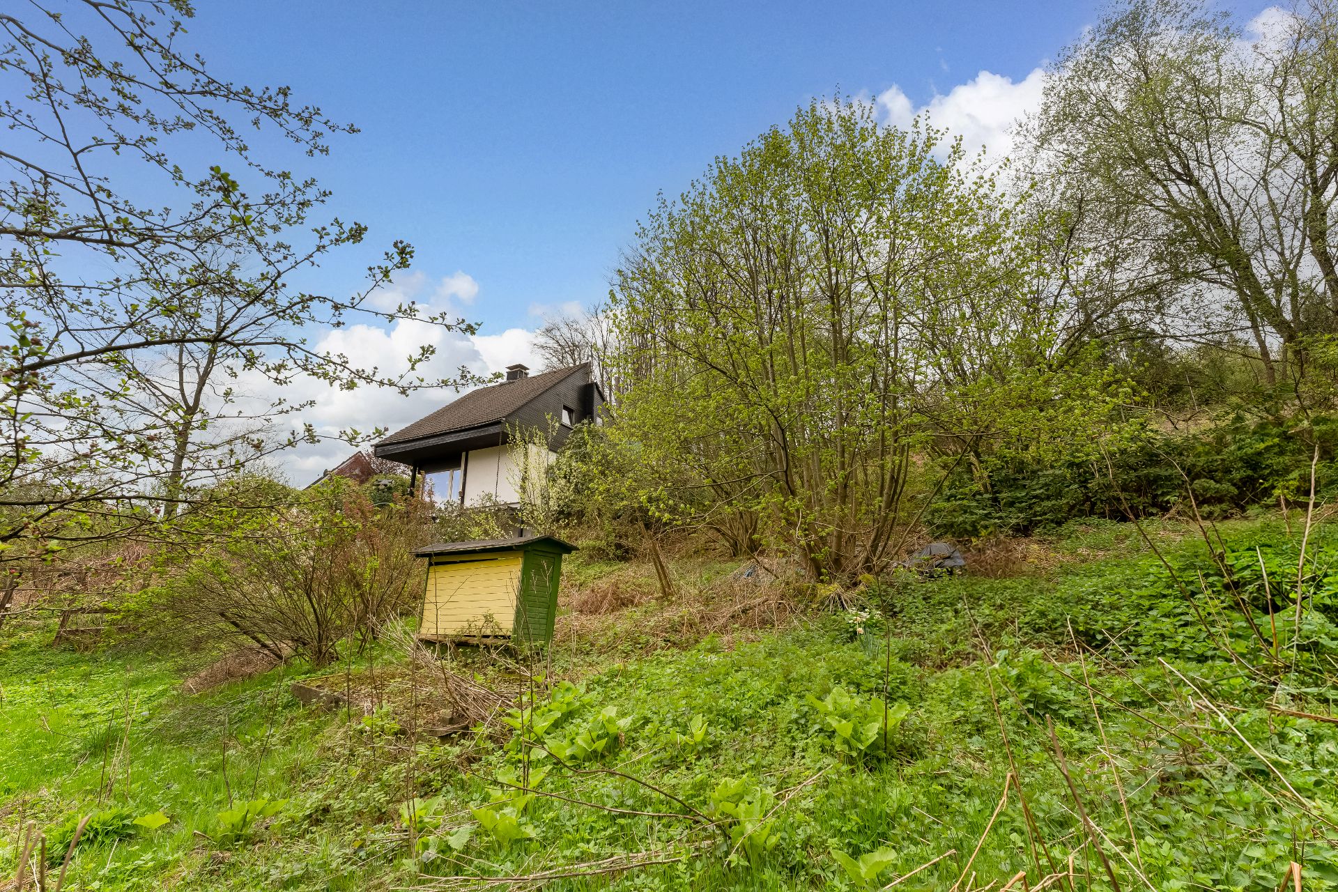 Haus mit dunklem Dach auf grünem Hang, Bäume und gelbes Gartenhaus im Vordergrund.