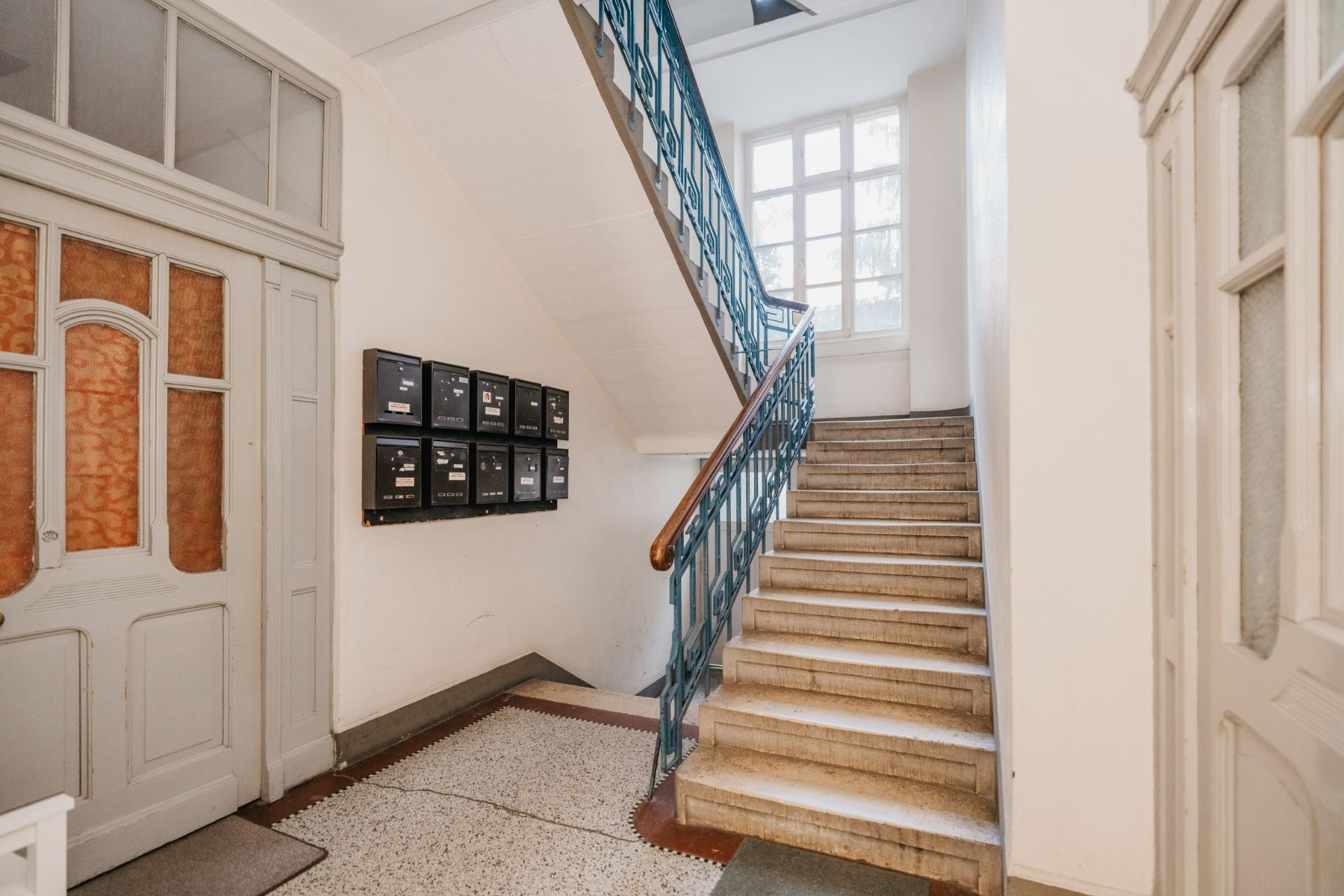 Helles Treppenhaus mit Briefkästen und blauem Geländer in einem Wohngebäude.