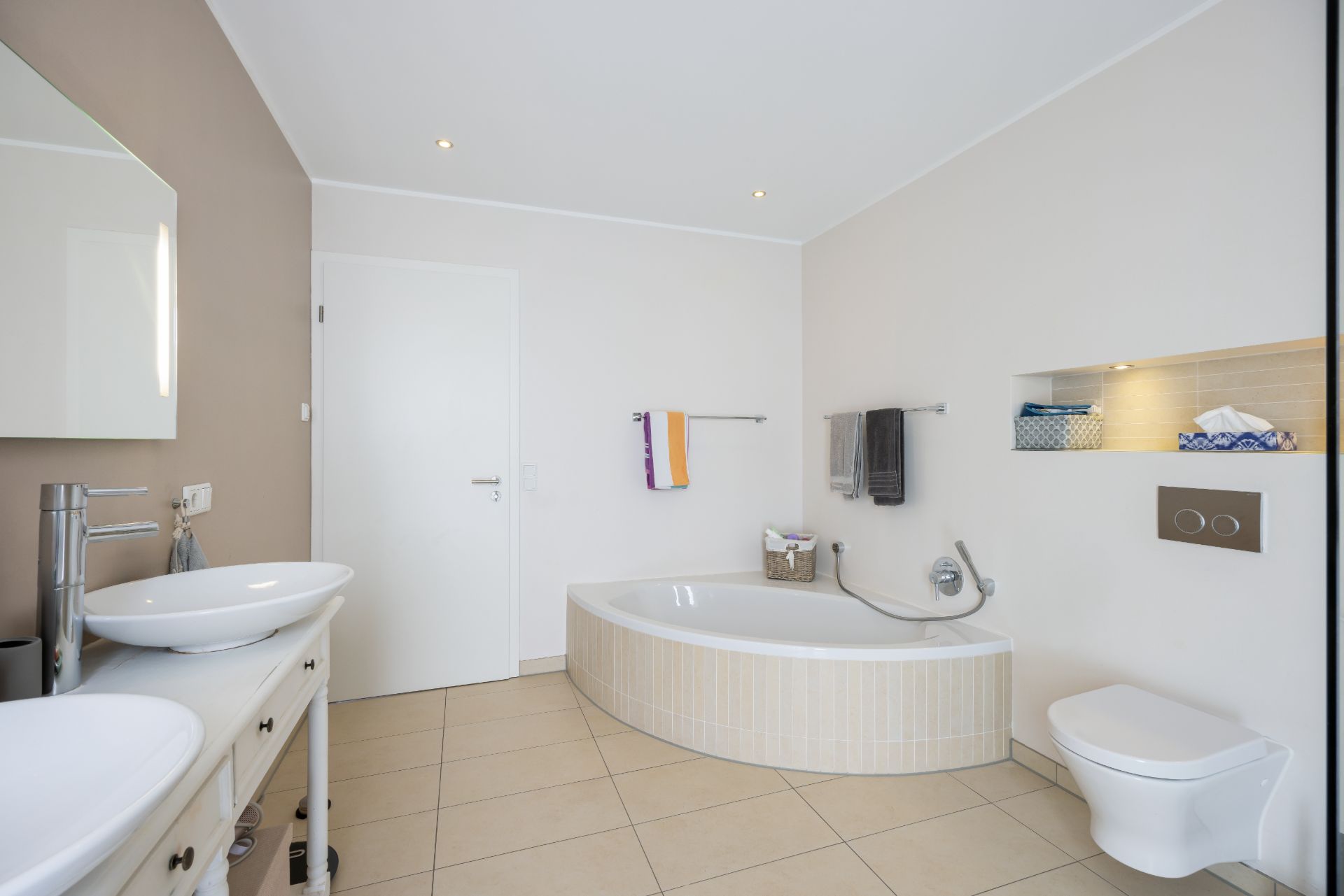 Modernes Badezimmer mit Eckbadewanne, zwei Waschbecken, WC und Handtüchern an den Wänden.
