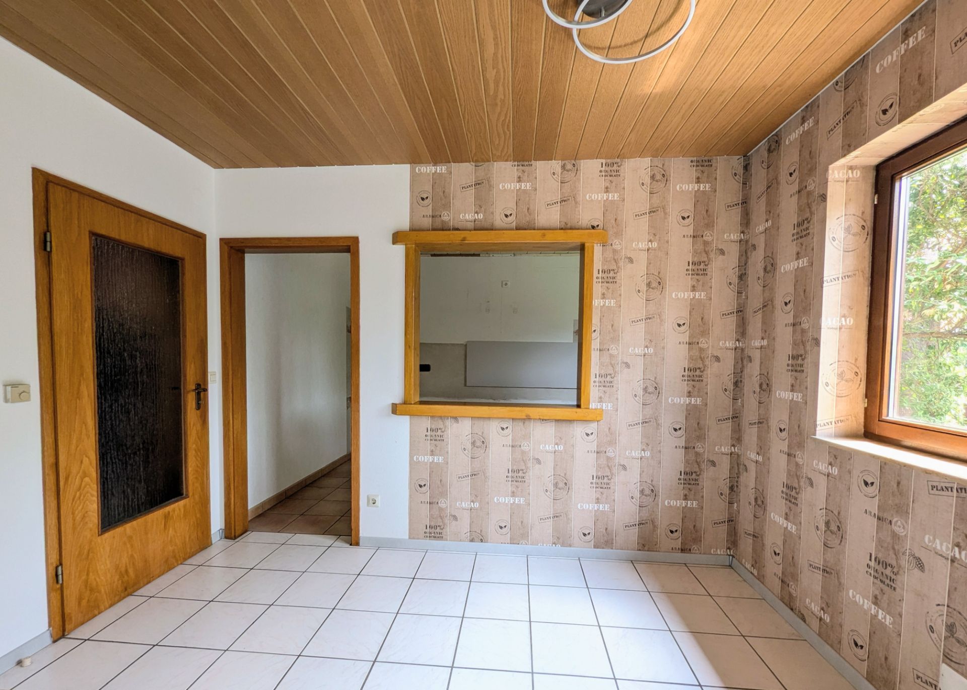 Heller Raum mit Fliesenboden, Holzdecke, Fenster, zwei Türen und Durchreiche zur nächsten Stube.