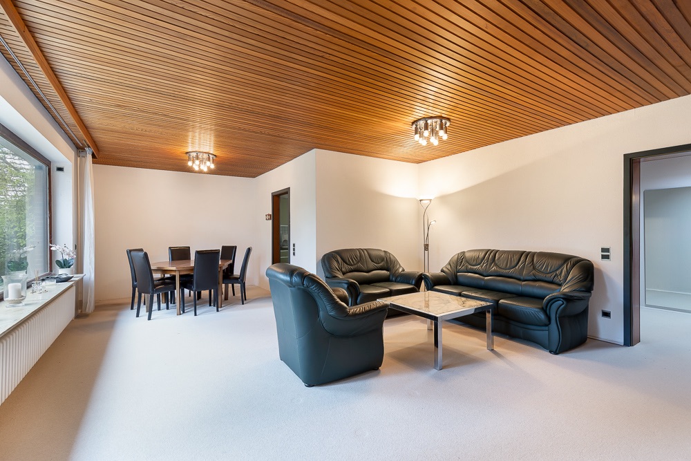 Wohnzimmer mit zwei schwarzen Ledersofas, Glastisch und Essbereich mit sechs Stühlen, Holzdecke.