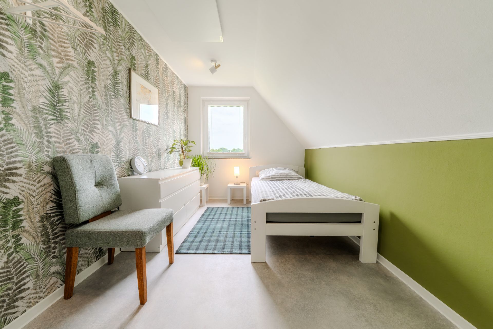 Helles Schlafzimmer mit Bett, Sessel, Kommode, Pflanze und grün gemusterter Tapete.