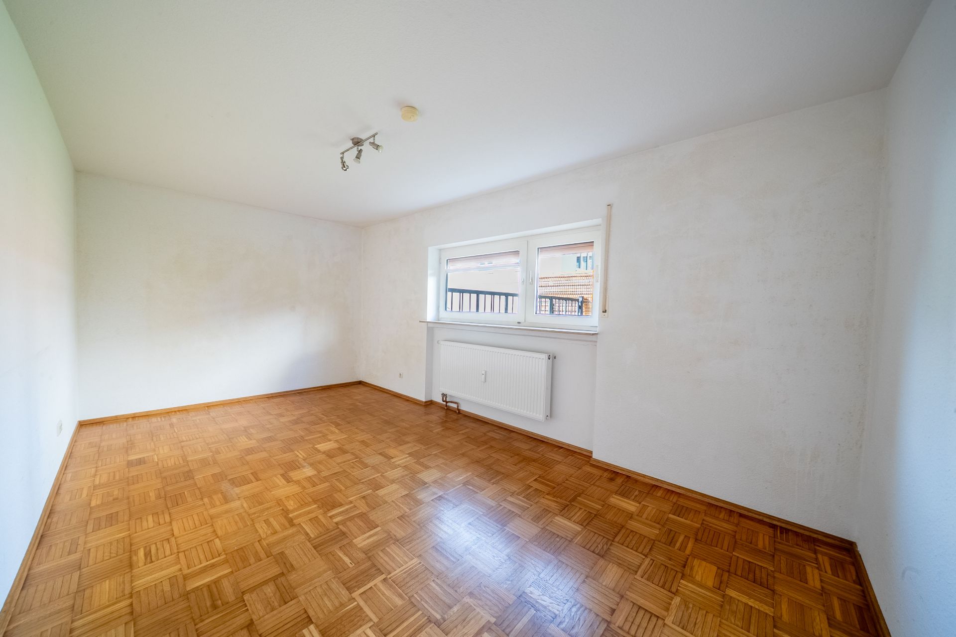 Leerer Raum mit Parkettboden, weiß gestrichenen Wänden und einem Fenster mit Heizung darunter.
