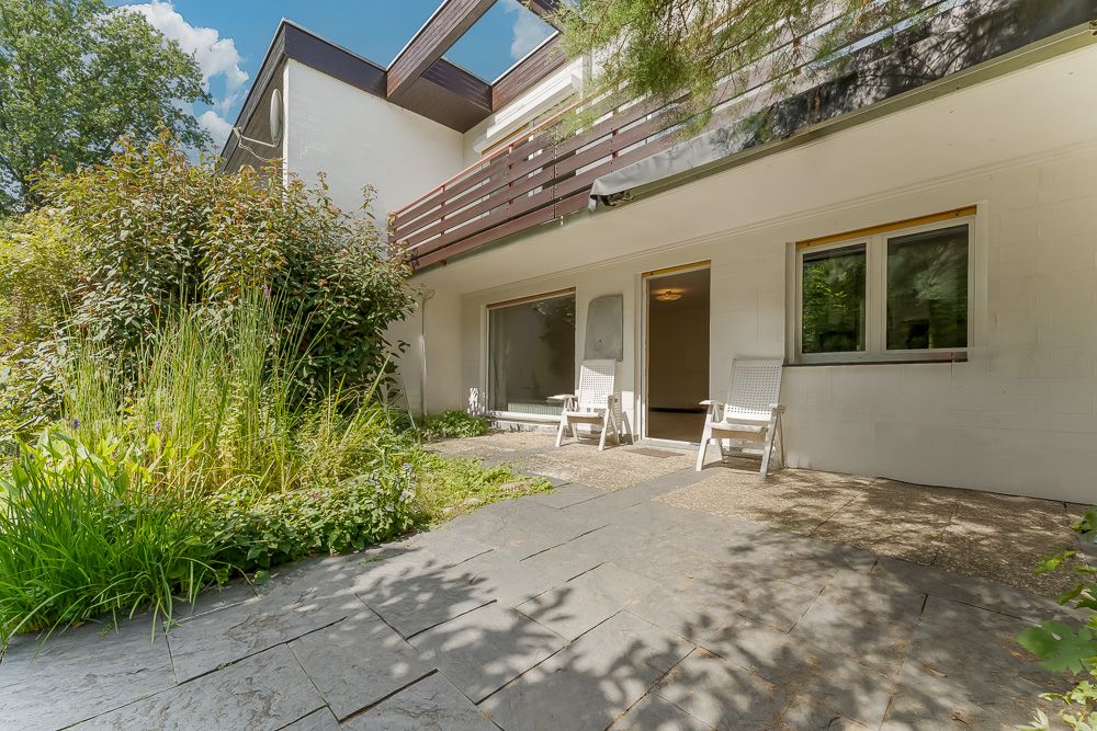 Steinterrasse mit zwei weißen Stühlen, davor Garten und moderne Hausfassade mit Balkon.