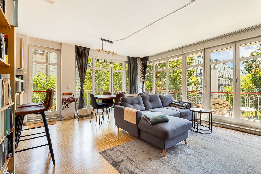 Helles Wohnzimmer mit großer Eckcouch, Essbereich und vielen Fenstern mit Blick auf Bäume.