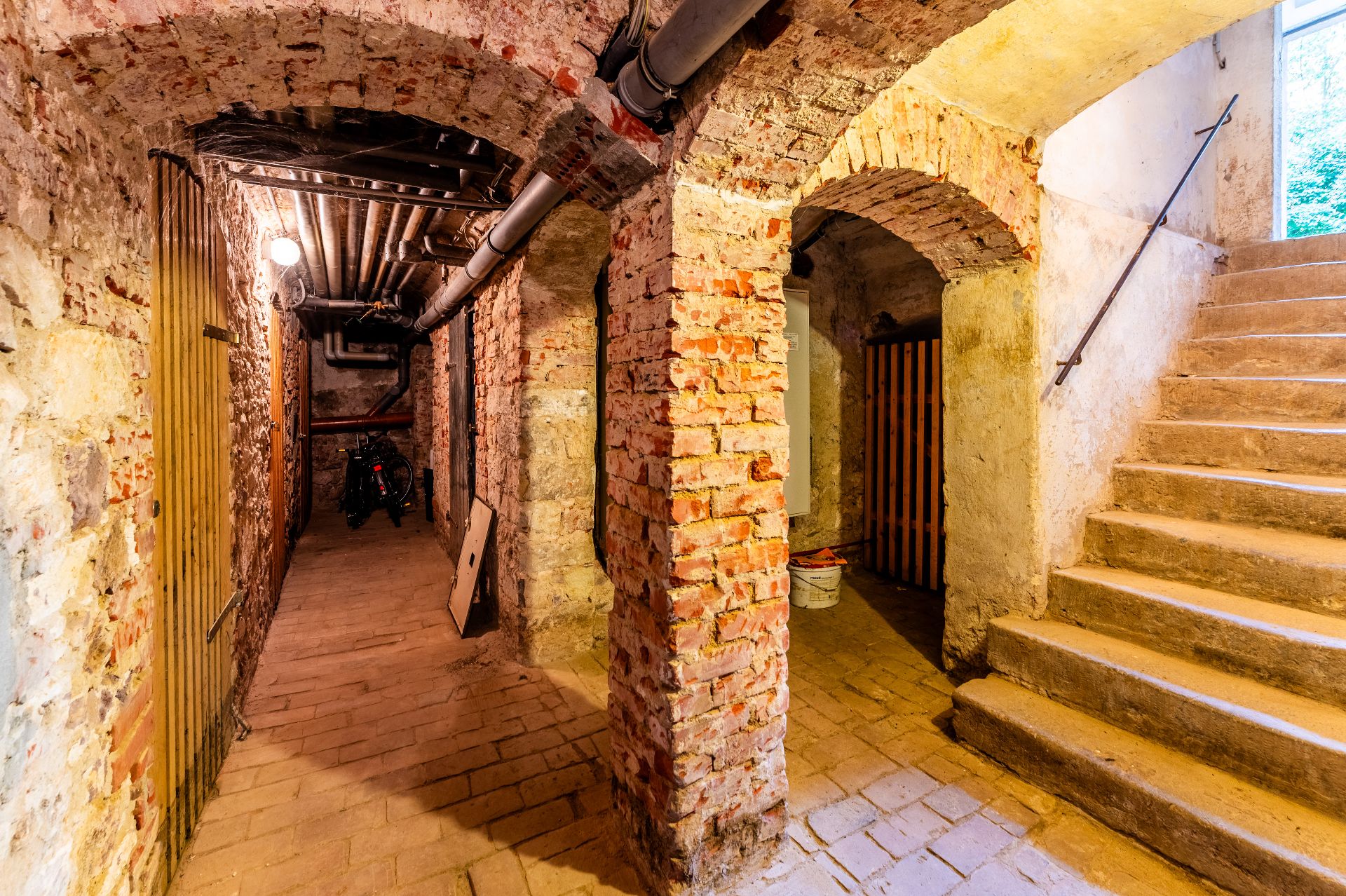 Alter gemauerter Keller mit Backsteinwänden, Gewölbebögen und einer Treppe nach oben.