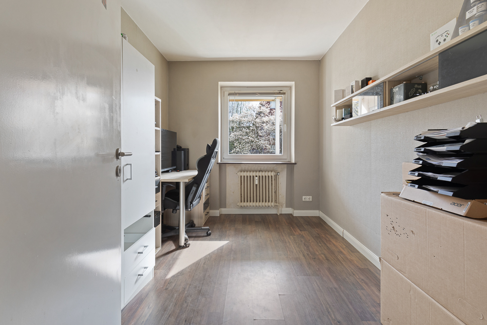 Helles Arbeitszimmer mit Schreibtisch, Computerstuhl, Regal und Fenster mit Blick nach draußen.