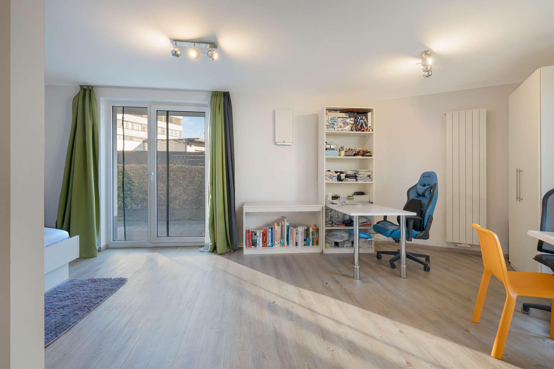 Helles Zimmer mit Schreibtischen, Regalen, Bürostühlen und Balkontür mit grünen Vorhängen.