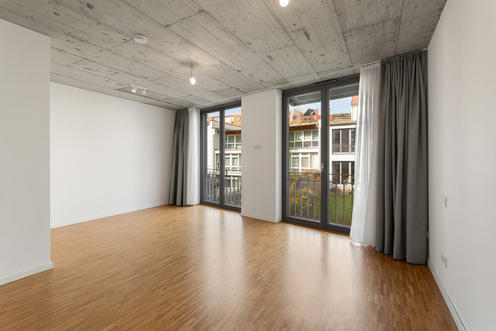 Leerer Raum mit Holzboden, Beton-Decke und großen Fenstern mit Vorhängen.