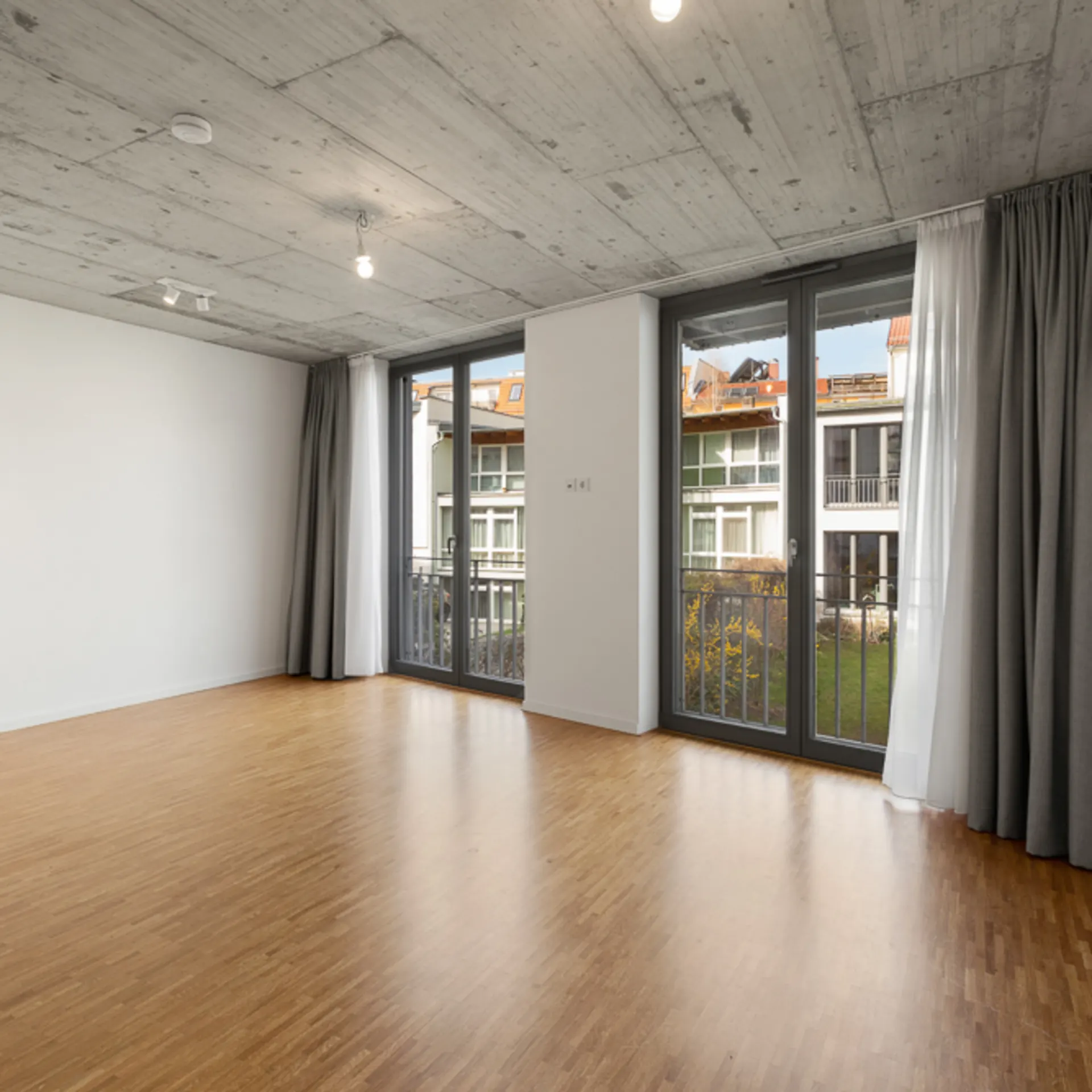 Leerer Raum mit Holzboden, Beton-Decke und großen Fenstern mit Vorhängen.