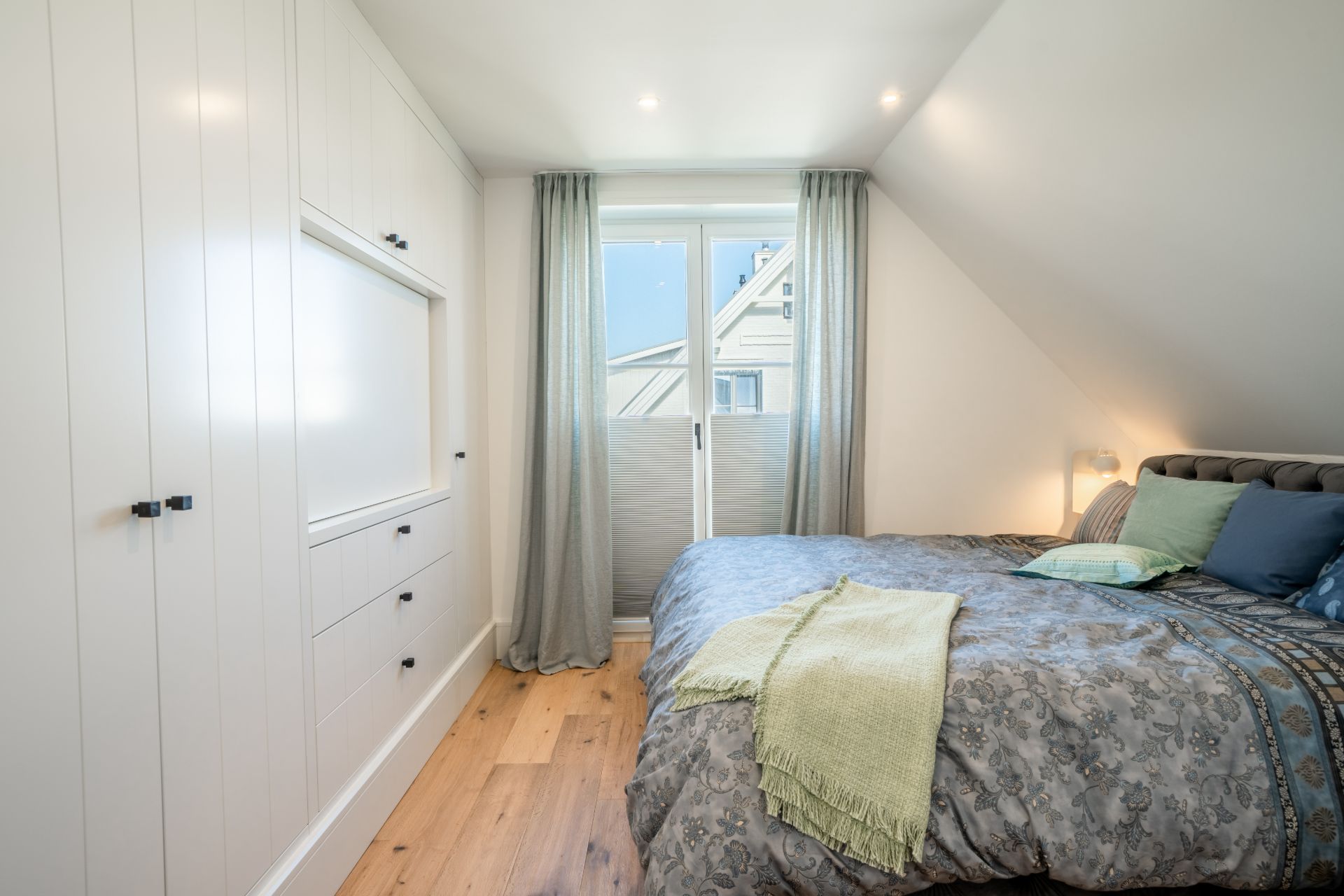 Helles Schlafzimmer mit Doppelbett, Einbauschrank und Fenster mit Vorhängen.