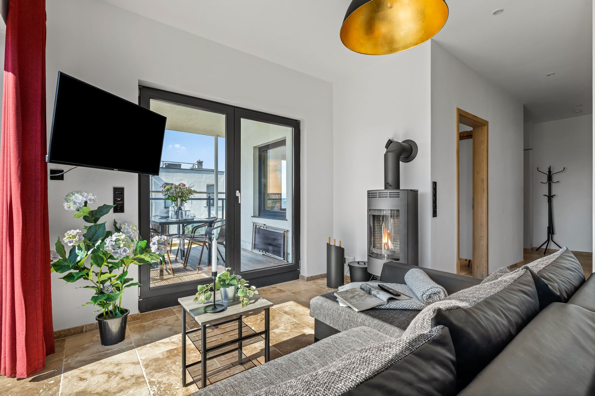 Wohnzimmer mit schwarzem Sofa, Kamin, Fernseher, Glastür zum Balkon und Zimmerpflanzen.