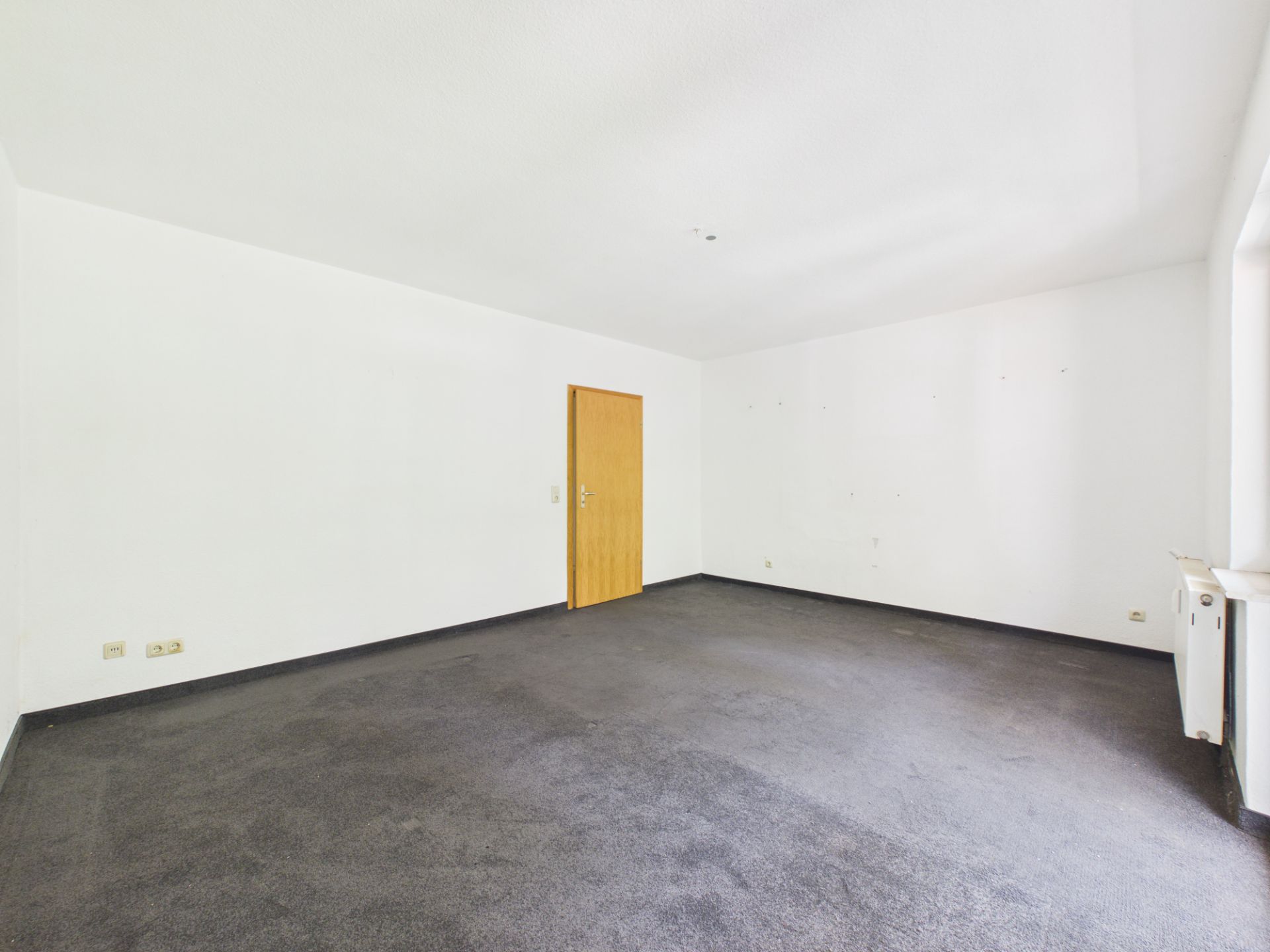 Leerer Raum mit weißer Wand, dunklem Teppichboden, Holztür und Fenster auf der rechten Seite.