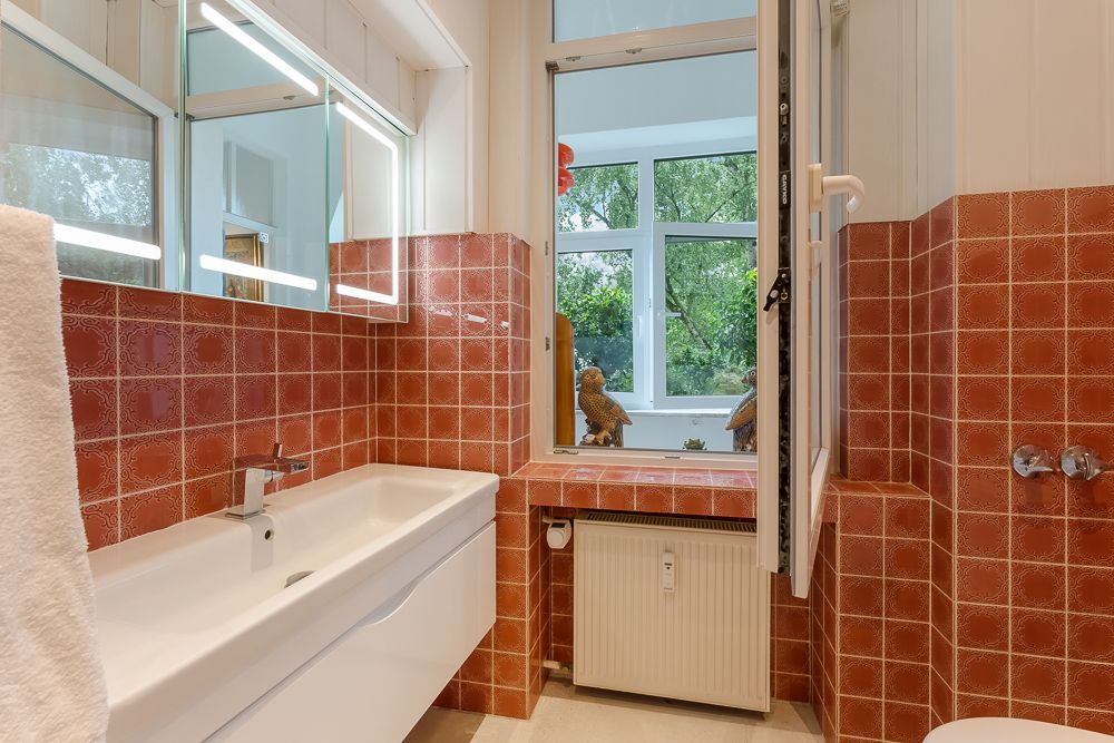 Badezimmer mit roten Fliesen, Waschbecken, Spiegel und offenem Fenster mit Blick ins Grüne.