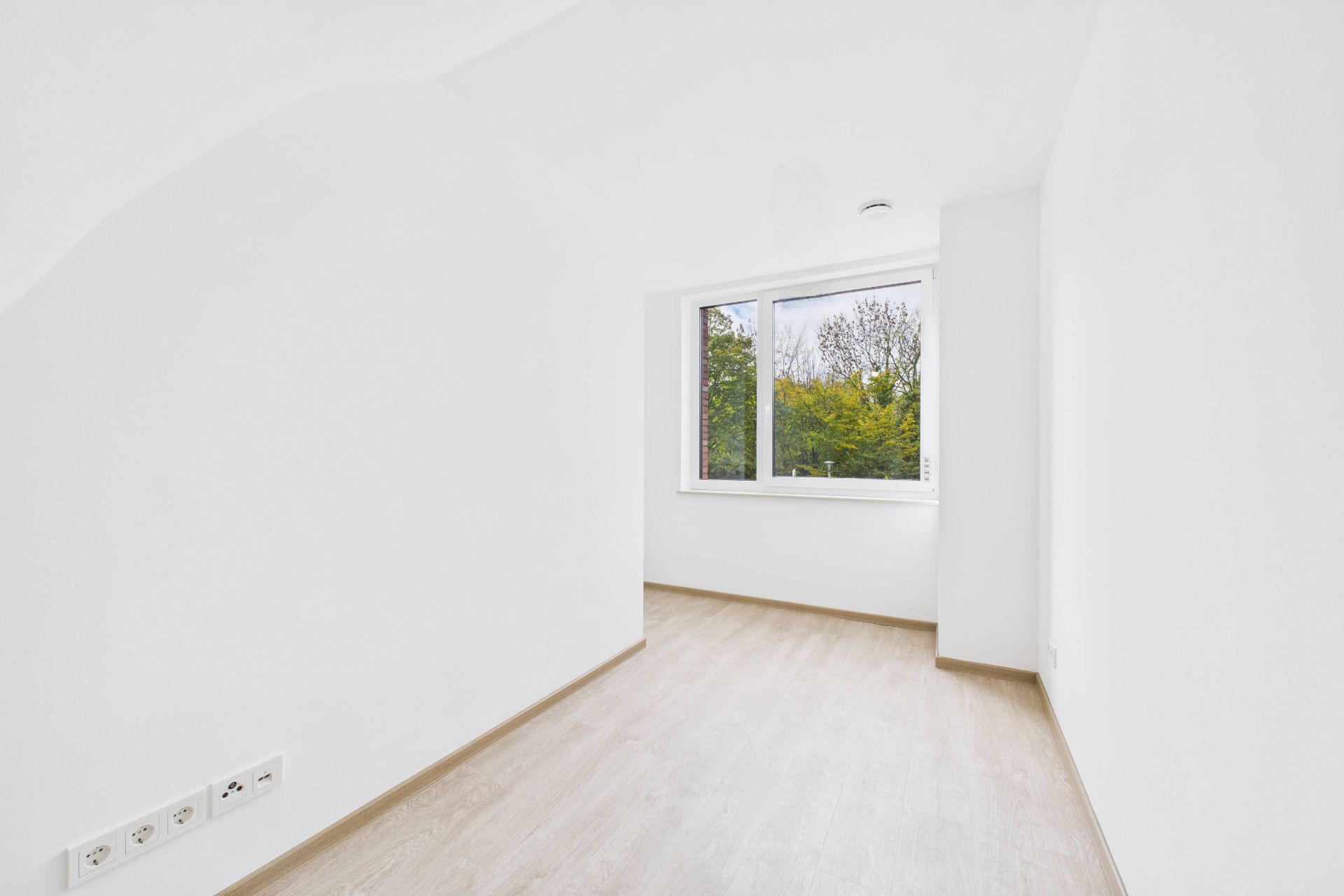 Heller, leerer Raum mit Holzfußboden und Fenster mit Blick ins Grüne.
