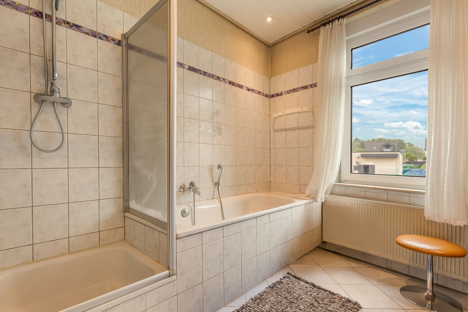 Gefliestes Badezimmer mit Badewanne, Dusche, Fenster mit Vorhang und Hocker.