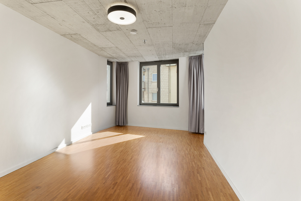 Leerer Raum mit Holzboden, weißen Wänden, Betondecke und zwei Fenstern mit Vorhängen.