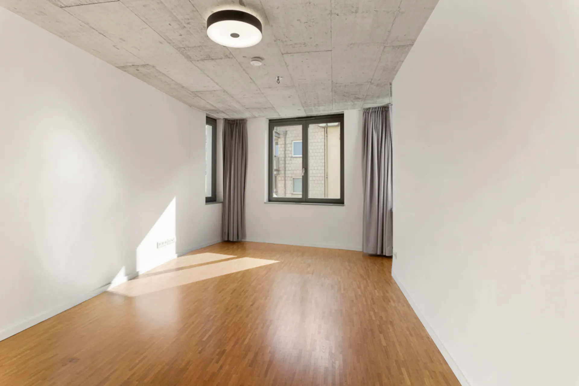 Leerer Raum mit Holzboden, weißen Wänden, Betondecke und zwei Fenstern mit Vorhängen.