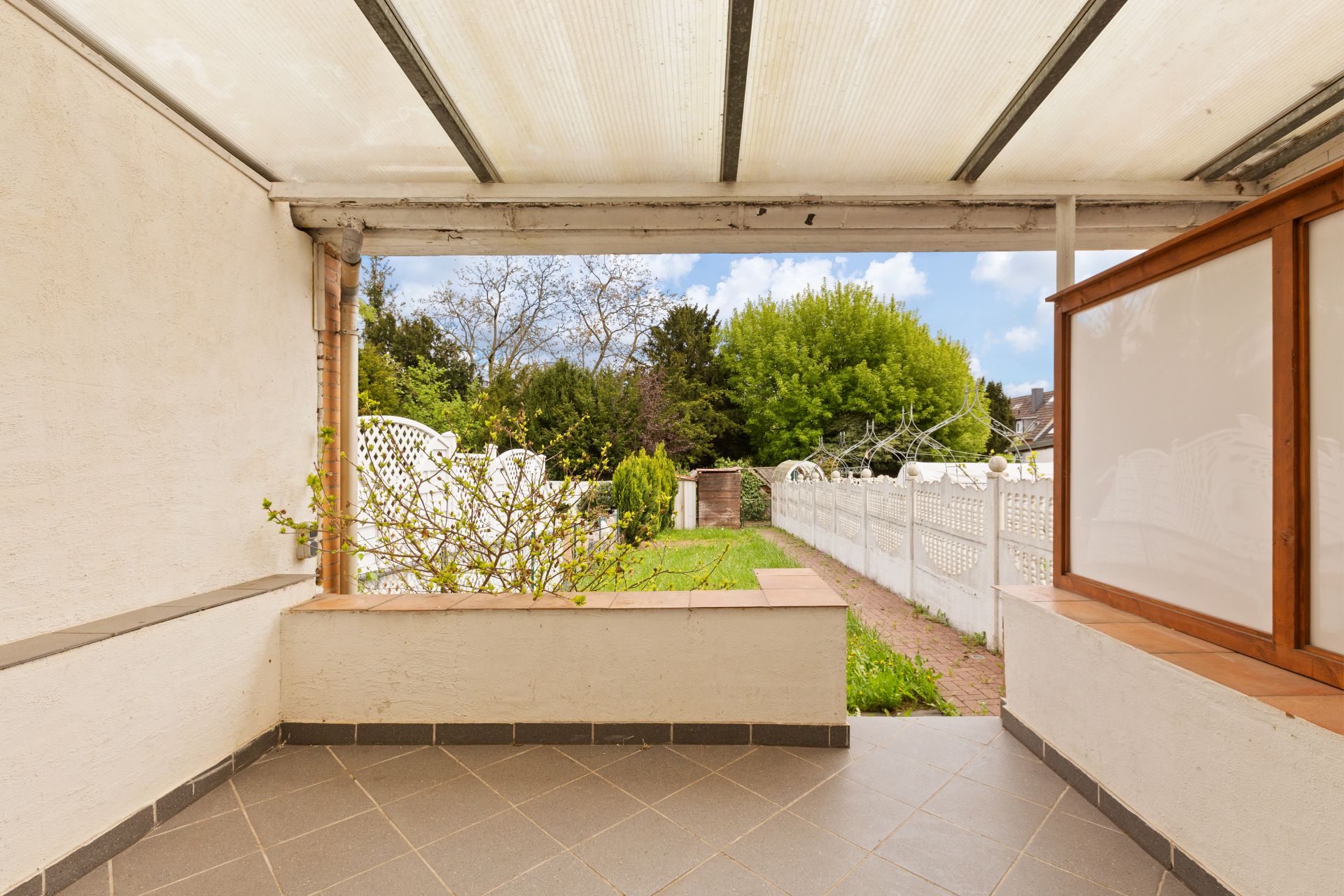 Überdachte Terrasse mit Fliesenboden, Blick auf Garten mit Rasen und weißen Zäunen.