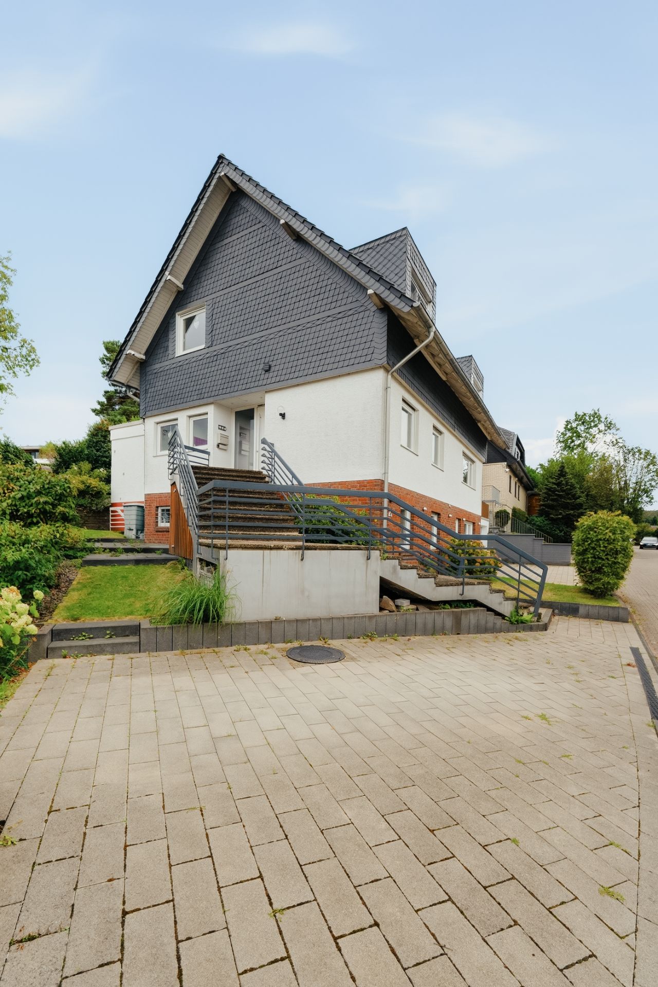 Modernes Wohnhaus mit dunkel gedecktem Dach, Treppe zum Eingang und gepflastertem Vorplatz.