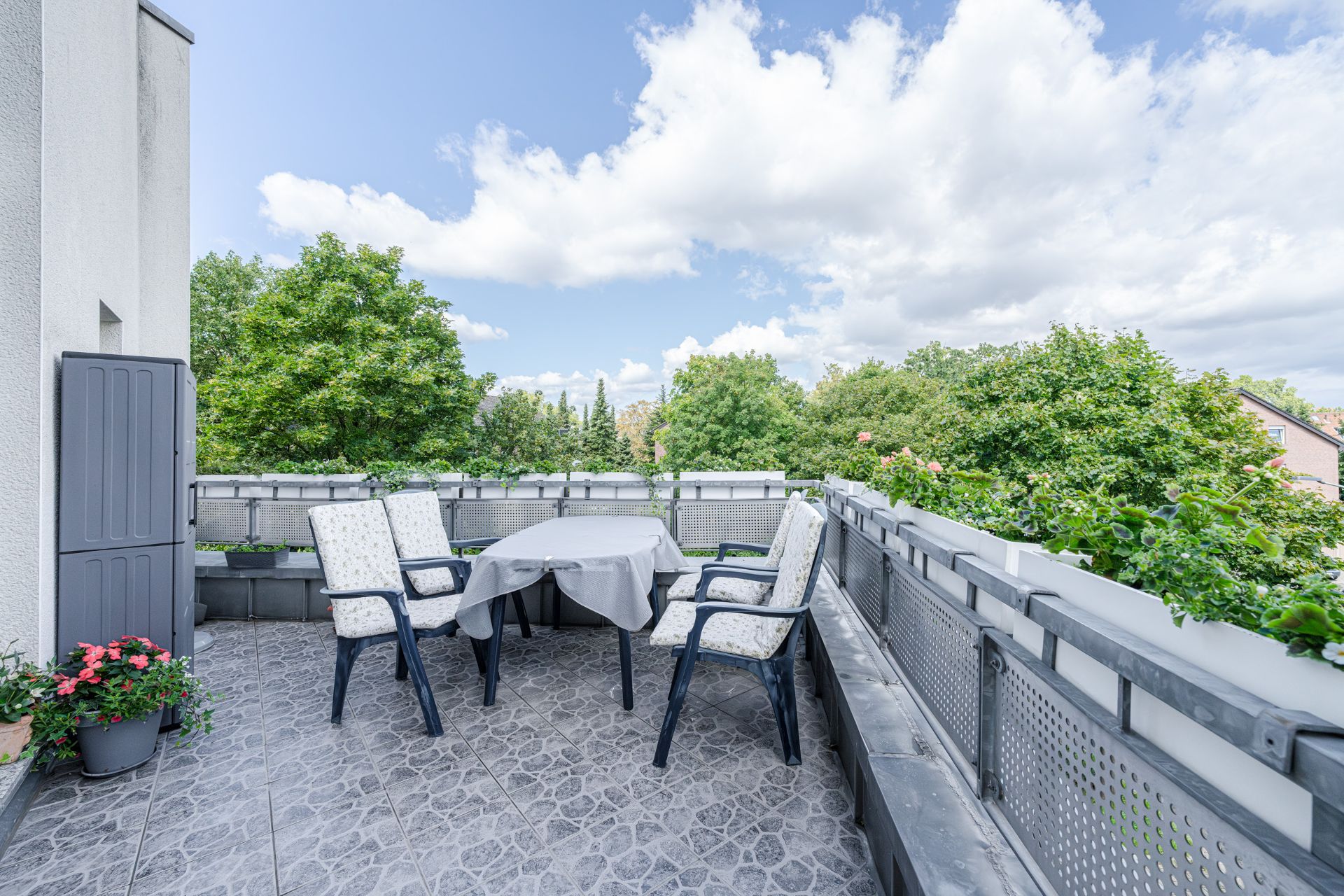 Große Terrasse mit vier Stühlen, Tisch und Pflanzen, umgeben von grünen Bäumen.