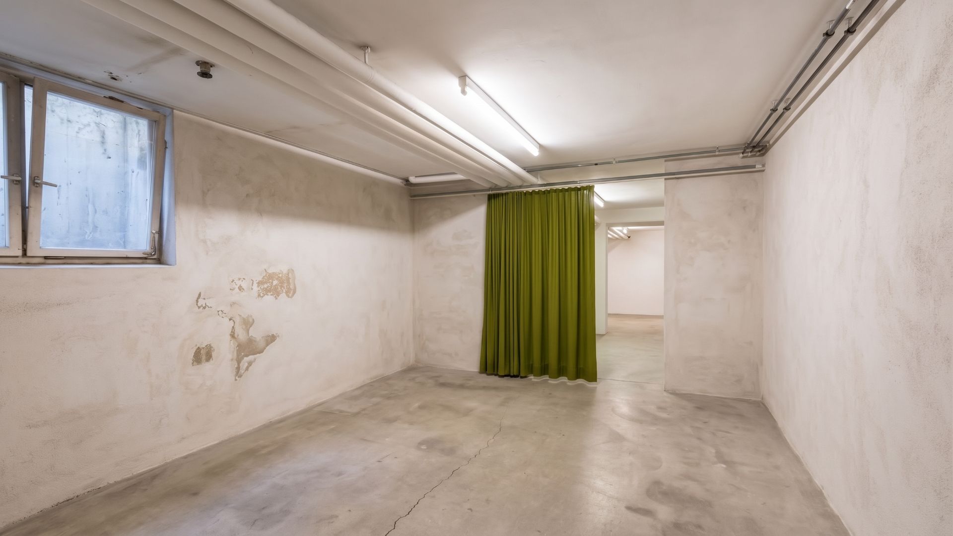 Kellerraum mit Betonboden, Fenster und grünem Vorhang an der Wand.