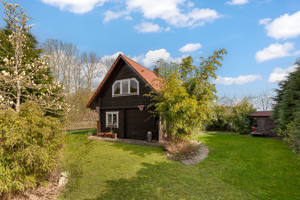 Braunes Holzhaus mit rotem Dach in grünem Garten, umgeben von Bäumen und Sträuchern.